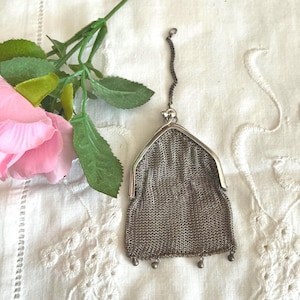 May include: An antique silver mesh purse with a chain handle and small decorative beads at the bottom. The purse is displayed on a white, embroidered cloth next to a pink rose and green leaves.
