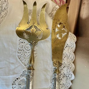 May include: Antique serving set featuring a gold-toned fork and knife. The fork has three tines and a decorative leaf design. The knife has a curved blade with an ornate handle. Both pieces have silver-toned handles with floral engravings.