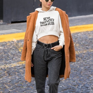 May include: A woman wearing a white cropped hoodie with the text "SEMI CRUNCHY MOMS CLUB" in black. She is also wearing a brown fuzzy coat, black jeans, and black sunglasses.