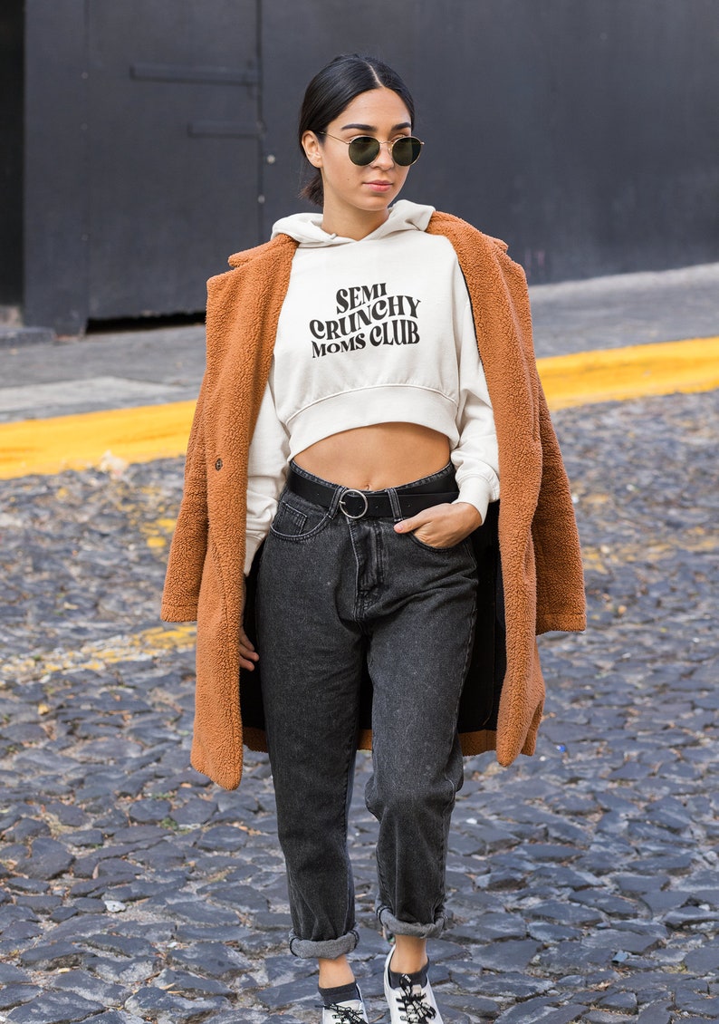 May include: A woman wearing a white cropped hoodie with the text "SEMI CRUNCHY MOMS CLUB" in black. She is also wearing a brown fuzzy coat, black jeans, and black sunglasses.