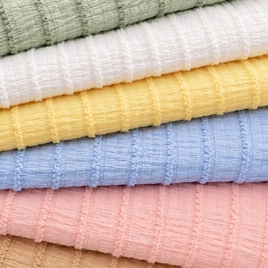Op de afbeelding: Close-up van een stapel textiel in verschillende pastelkleuren. De kleuren zijn lichtgroen, wit, geel, lichtblauw, roze en bruin. De stof heeft een geribbeld patroon.