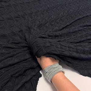 May include: Black fabric with a textured, quilted pattern. The fabric is being held by a hand wearing a silver bracelet.