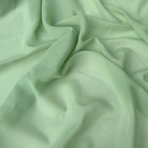 以下が含まれることがあります： 柔らかく流れるような質感の薄い緑色のシアー生地。生地はドレープされ、折り畳まれ、繊細で通気性の良い品質が示されています。