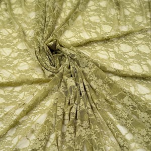 May include: Olive green lace fabric with a floral pattern. The fabric is draped and folded, creating shadows and depth. The lace has a delicate, open weave, and the color is a muted, earthy tone. This fabric is suitable for apparel and craft projects.
