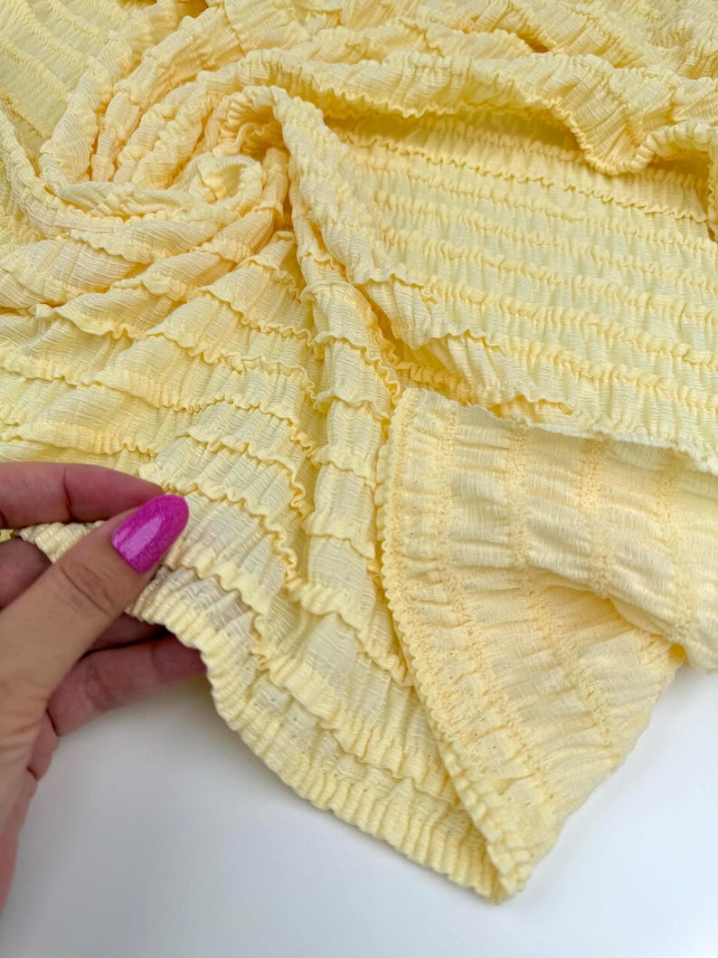 Op de afbeelding: Close-up van een lichtgele stof met een getextureerd, gerimpeld ontwerp. De stof is verzameld en gevouwen, met de gedetailleerde, horizontale rijen van het materiaal. De kleur is een zacht, pastelgeel.