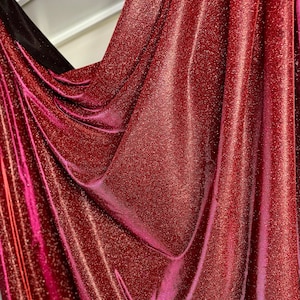 May include: Red fabric with a sparkly, glittery texture. The fabric is draped and folded, showing the shimmery effect of the material.