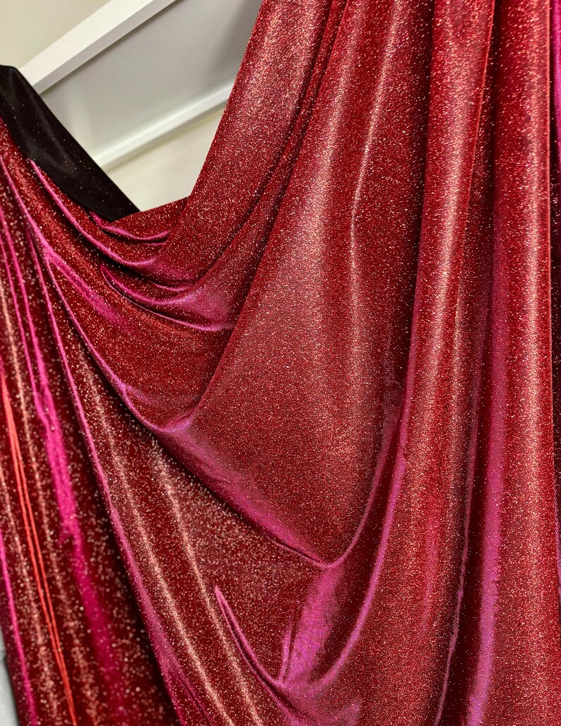 May include: Red fabric with a sparkly, glittery texture. The fabric is draped and folded, showing the shimmery effect of the material.