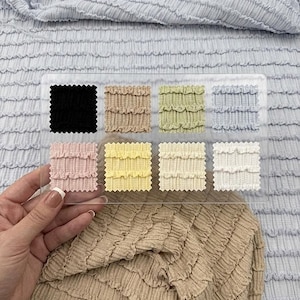 Op de afbeelding: Een hand houdt een doorzichtige acryl stalenkaart met stofstalen in verschillende kleuren. De stalen omvatten zwart, beige, groen, blauw, roze, geel, cr&egrave;me en wit. De achtergrond toont lichtblauwe en beige getextureerde stof.