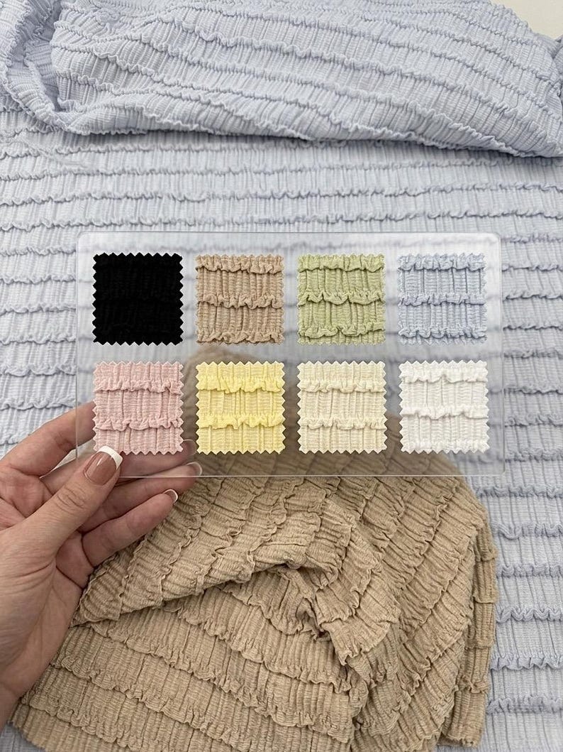 Op de afbeelding: Een hand houdt een doorzichtige acryl stalenkaart met stofstalen in verschillende kleuren. De stalen omvatten zwart, beige, groen, blauw, roze, geel, cr&egrave;me en wit. De achtergrond toont lichtblauwe en beige getextureerde stof.
