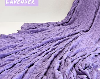 May include: A close-up of a textured, lavender-colored fabric. The fabric has a crinkled, quilted appearance. The word "LAVENDER" is written in white on a purple background in the top left corner of the image.