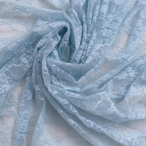May include: Baby blue lace fabric with a floral pattern, draped and folded. The fabric is a light, airy material, with the words "BABY BLUE" in a white box at the bottom.