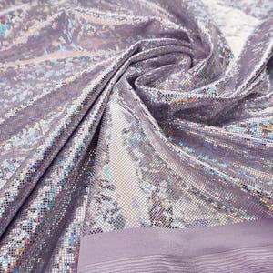 May include: A close-up of a shimmering, iridescent fabric in shades of lavender and silver. The material is heavily textured with a reflective, sequin-like pattern. The fabric is folded, revealing a solid lavender border with horizontal lines.