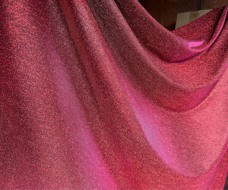 May include: A close-up of a red fabric with a sparkly, glittery texture. The fabric is draped and folded, creating soft, flowing lines.