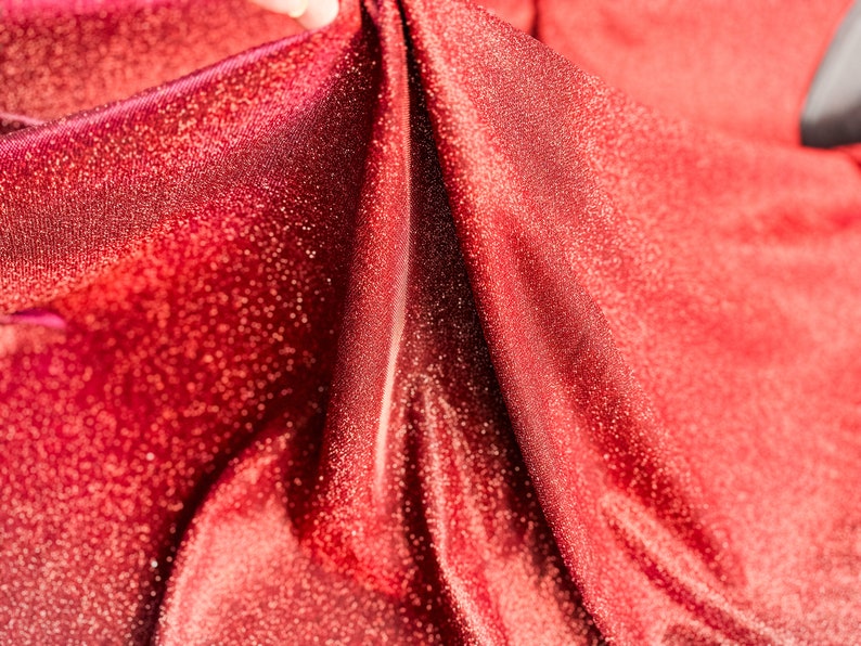 May include: Close-up of red fabric with a sparkly texture. The fabric is folded and draped, creating a soft, flowing look.