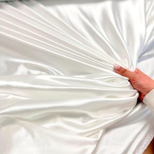 K&ouml;nnte beinhalten: Wei&szlig;er Satinstoff mit einer glatten, seidenartigen Textur. Der Stoff ist gefaltet und drapiert und zeigt seine luxuri&ouml;se Haptik.