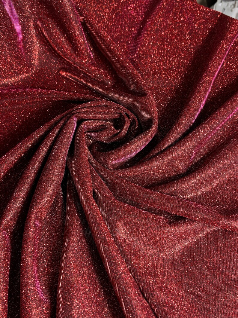 May include: Red sparkly fabric with a shimmery finish. The fabric is folded and draped, showing its texture and sheen.