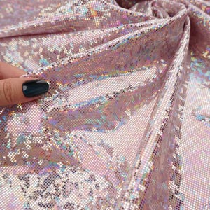 May include: Close-up of a pink, iridescent fabric with a shimmering, textured surface. The fabric is folded, revealing multiple layers and a subtle, grid-like pattern. The material has a holographic effect, reflecting various colors.