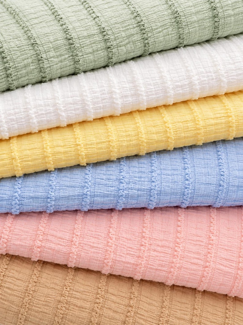Op de afbeelding: Close-up van een stapel textiel in verschillende pastelkleuren. De kleuren zijn lichtgroen, wit, geel, lichtblauw, roze en bruin. De stof heeft een geribbeld patroon.