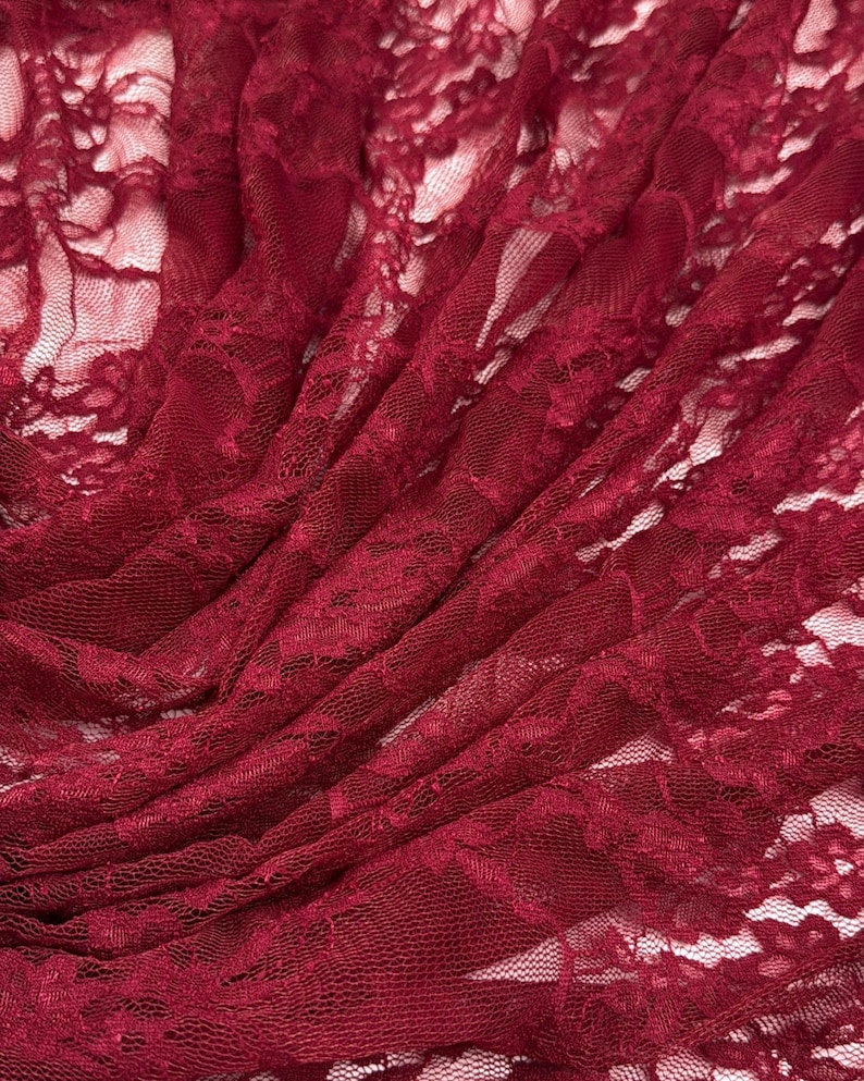 K&ouml;nnte beinhalten: Nahaufnahme eines tiefroten Spitzenstoffs. Das zarte Blumenmuster ist im gesamten Material sichtbar. Der Stoff ist drapiert, wodurch Schatten entstehen und die Textur hervorgehoben wird. Die Spitze wird wahrscheinlich f&uuml;r Kleidung oder Bastelprojekte verwendet.