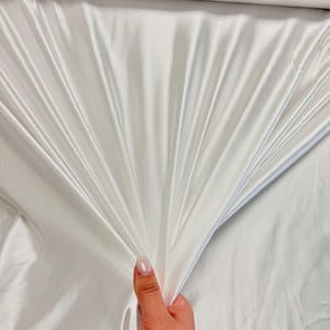K&ouml;nnte beinhalten: Wei&szlig;er Satin-Stoff mit einer glatten, seidenartigen Textur. Der Stoff ist drapiert und gefaltet und zeigt seine luxuri&ouml;se Haptik.