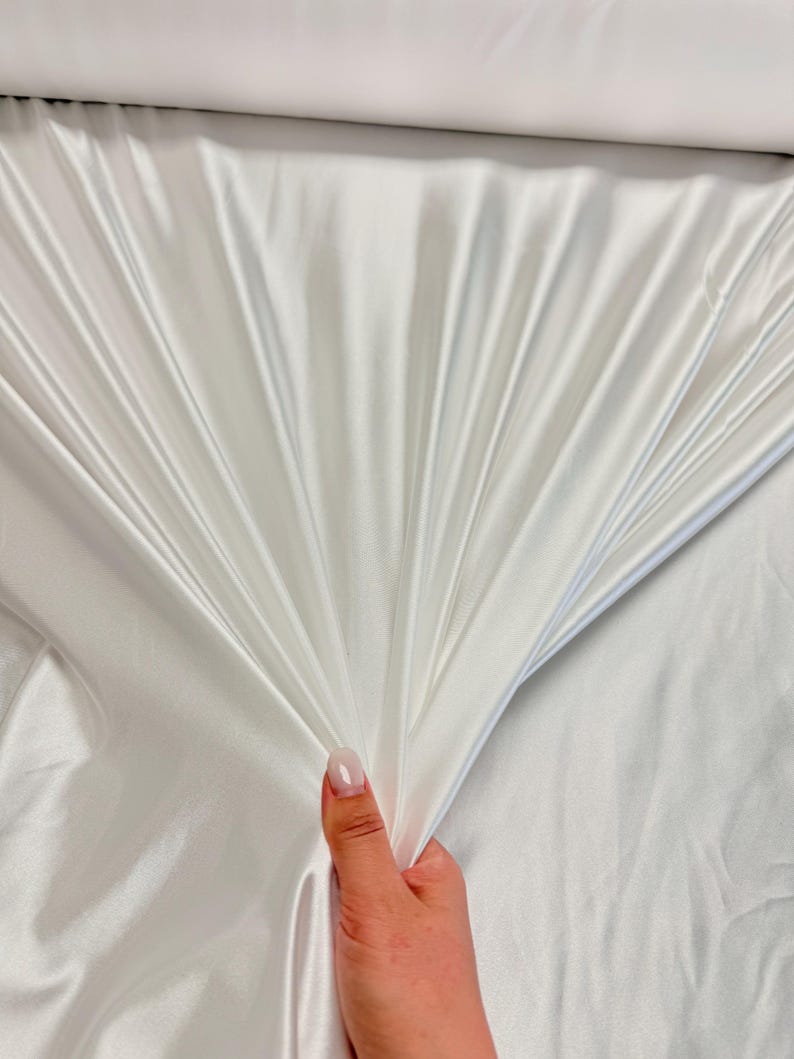 K&ouml;nnte beinhalten: Wei&szlig;er Satin-Stoff mit einer glatten, seidenartigen Textur. Der Stoff ist drapiert und gefaltet und zeigt seine luxuri&ouml;se Haptik.