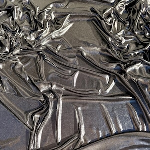 May include: A dark charcoal grey fabric with a metallic, reflective finish. The material is draped in deep, irregular folds that create high-contrast highlights and shadows across the surface, emphasising the smooth, shimmering texture of the cloth.