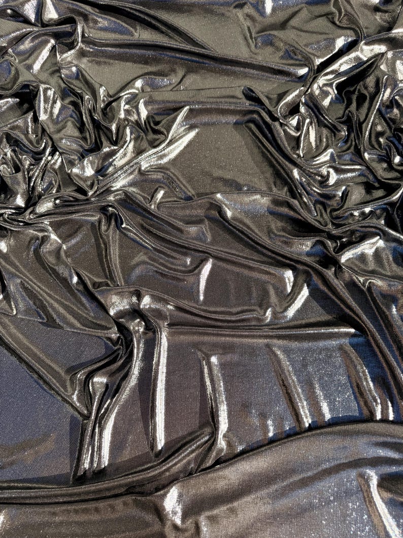 May include: A dark charcoal grey fabric with a metallic, reflective finish. The material is draped in deep, irregular folds that create high-contrast highlights and shadows across the surface, emphasising the smooth, shimmering texture of the cloth.
