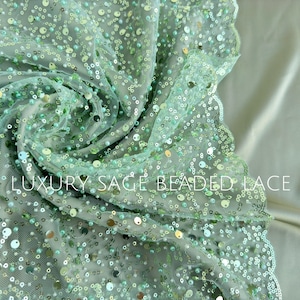 May include: Sage green beaded lace fabric with a scalloped edge. The fabric is made of a fine mesh with small, round beads scattered throughout.