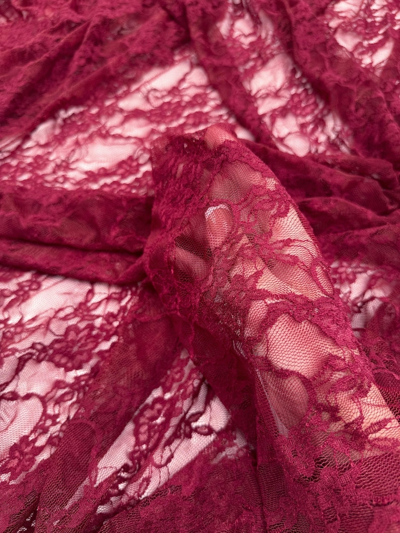 K&ouml;nnte beinhalten: Nahaufnahme eines bordeauxfarbenen Spitzenstoffs. Das zarte Blumenmuster ist im gesamten transparenten Material sichtbar. Der Stoff ist gerafft, wodurch Falten und Schatten entstehen, die die Textur und Tiefe der Spitze hervorheben. Die Farbe ist ein tiefer, satter Rotton.