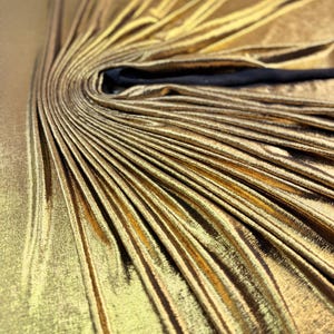 May include: A metallic gold fabric garment featuring a dense, sunburst-style pleated design radiating from a central point. The material has a reflective, shimmering finish with deep folds and a smooth texture, accented by a solid black strap or trim.
