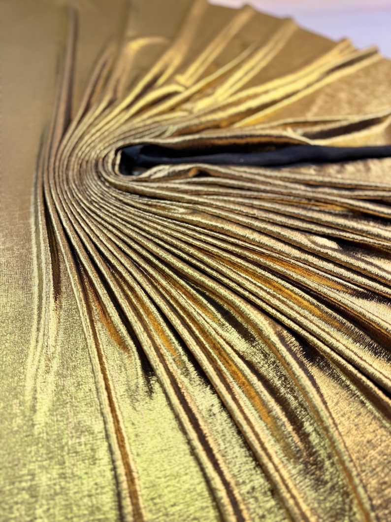 May include: A metallic gold fabric garment featuring a dense, sunburst-style pleated design radiating from a central point. The material has a reflective, shimmering finish with deep folds and a smooth texture, accented by a solid black strap or trim.