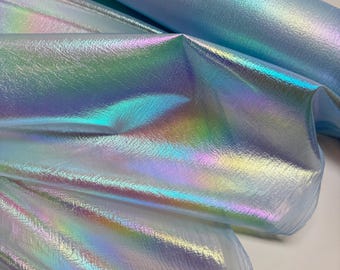May include: Iridescent fabric in shades of blue, green, and purple. The material has a textured surface and a shimmering, holographic effect. Rolls of the fabric are visible, with some sections draped to show the color variations.