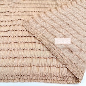 Op de afbeelding: Taupe stof met een getextureerd, gerimpeld ontwerp. De stof is dubbelgevouwen, wat de textuur en kleur laat zien. De tekst "Taupe" is zichtbaar, samen met de tekst "Multiple yards will come in one piece."
