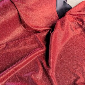 May include: Close-up of a red fabric with a sparkly, iridescent finish. The fabric is folded and draped, showing its texture and sheen.
