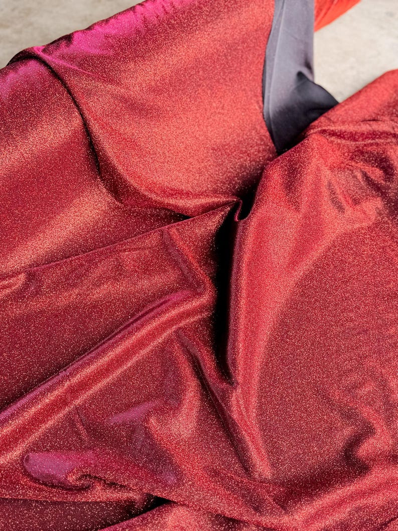 May include: Close-up of a red fabric with a sparkly, iridescent finish. The fabric is folded and draped, showing its texture and sheen.