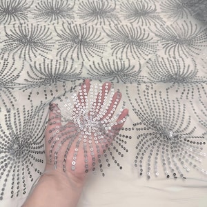 May include: Silver sequin fabric with a repeating floral pattern. The sequins are arranged in a radial design, creating a shimmering effect. The fabric is a light color, possibly white or off-white, and the sequins are a metallic silver.