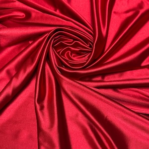 Può includere: Primo piano di tessuto di raso rosso vibrante. Il tessuto è drappeggiato e arricciato con cura, creando un effetto a spirale al centro. Il materiale liscio e lucido riflette la luce, evidenziando le pieghe e le trame del tessuto.