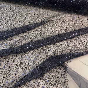 May include: Close-up of navy blue fabric covered in small, shimmering sequins. The fabric is draped, creating folds and shadows that highlight the texture and sparkle of the sequins. The overall effect is a luxurious, elegant look.