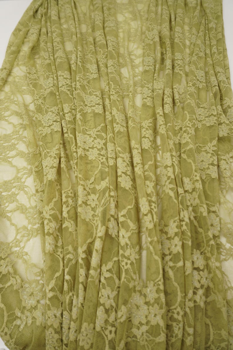 Puede incluir: Primer plano de una tela de encaje verde claro con un estampado floral. El delicado encaje est&aacute; drapeado, creando pliegues y sombras suaves. La tela tiene una calidad ligeramente transparente, revelando un vistazo del fondo.