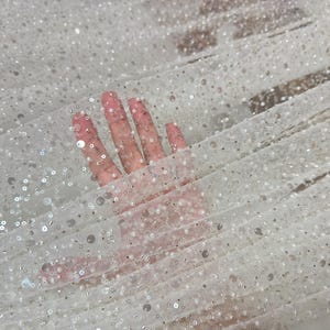 Könnte beinhalten: Weißer Tüllstoff mit kleinen, silbernen Pailletten, die über den Stoff verteilt sind. Der Stoff hat eine zarte, schimmernde Optik.