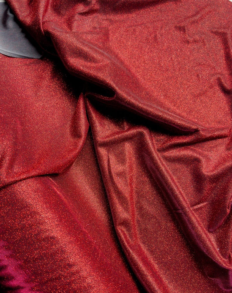 May include: Red sparkly fabric with a textured, glittery surface. The fabric is folded and draped, showing the shimmery texture.