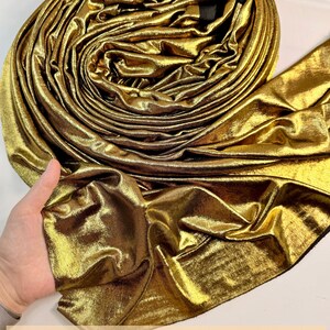 May include: A roll of metallic gold fabric with a shimmering, reflective surface and a soft, draped texture. The fabric is coiled into a circular shape, showcasing its bright, lustrous finish. Text at the bottom reads: ORDER MULTIPLE YARDS, RECEIVE IN ONE CONTINUOUS PIECE.