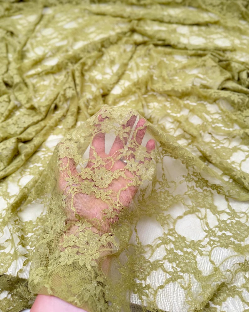 May include: Close-up of olive green lace fabric. The delicate floral pattern is visible throughout the sheer material. The fabric is draped, creating soft folds and shadows. The lace is held up, allowing light to pass through.