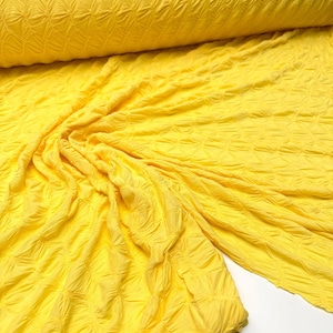 May include: Bright yellow fabric with a textured, quilted pattern. The fabric is draped and folded, revealing the texture and color variations. A roll of the same fabric is visible in the background.