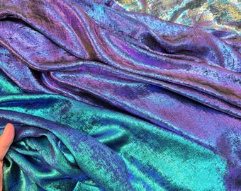 May include: Close-up of iridescent fabric in shades of teal, purple, and silver. The fabric has a shimmering, textured appearance with visible folds and creases, creating a dynamic visual effect. The material appears to be soft and flowing.