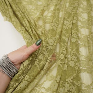 May include: Close-up of olive green lace fabric with a floral pattern. The delicate material is draped, revealing the intricate details of the design. The fabric is held by a hand with a silver bracelet.