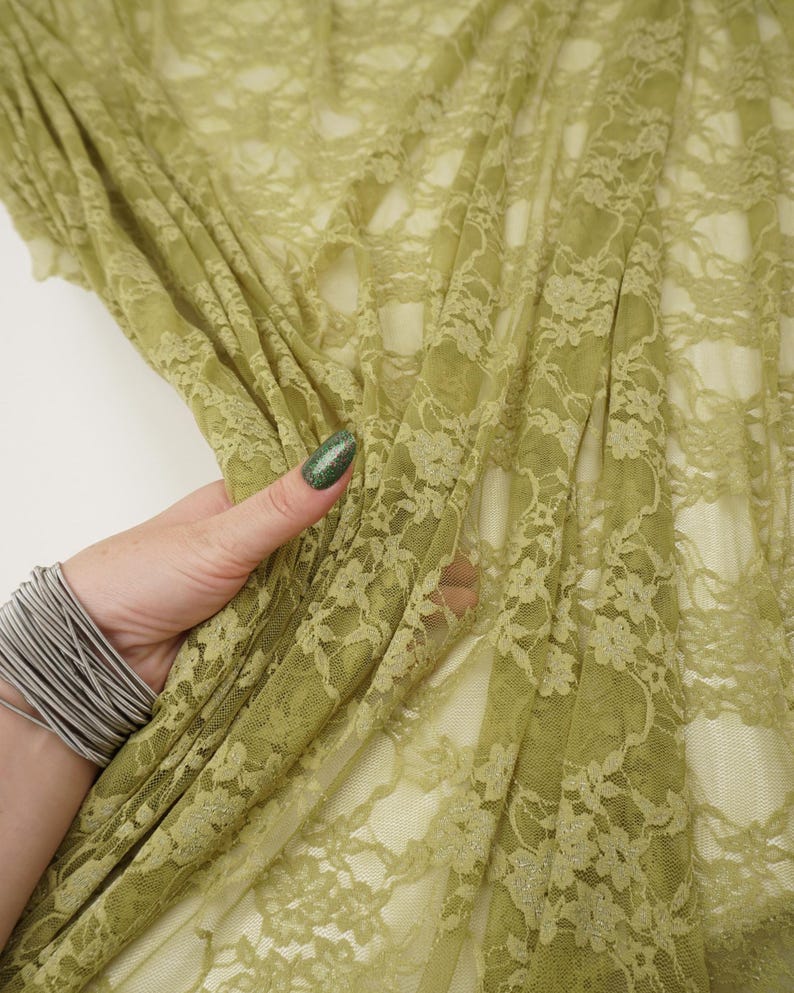Close-up of olive green lace fabric with a floral pattern. The delicate material is draped, revealing the intricate design. The fabric is held in place by a hand wearing a silver bracelet.