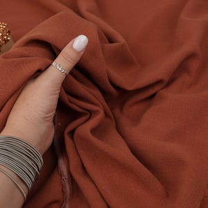 May include: A piece of solid, rust-colored fabric with a visible woven texture. The material is draped in soft folds, showcasing its matte finish and consistent deep orange-brown hue across the entire surface of the textile material.