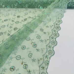 Peut inclure: Gros plan sur un tissu en tulle vert clair avec broderies florales et paillettes iris&eacute;es. Le tissu est enroul&eacute; &agrave; l'arri&egrave;re-plan et pr&eacute;sente un bord festonn&eacute;. La broderie comprend des fleurs et des &eacute;toiles.