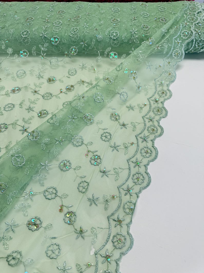 Peut inclure: Gros plan sur un tissu en tulle vert clair avec broderies florales et paillettes iris&eacute;es. Le tissu est enroul&eacute; &agrave; l'arri&egrave;re-plan et pr&eacute;sente un bord festonn&eacute;. La broderie comprend des fleurs et des &eacute;toiles.